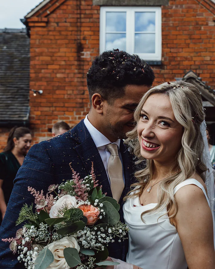 Bride smiling at the camera as her groom leans in, guests behind