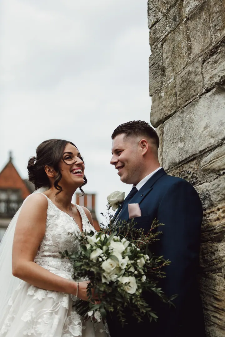 A bride laughing as her groom leans on an old stone wall at Hoar Cross Hall