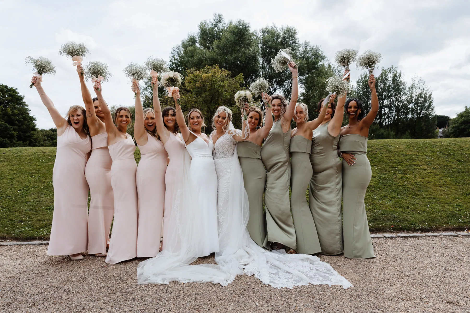 Two bridal parties cheering with their bouquets on a riverside lawn